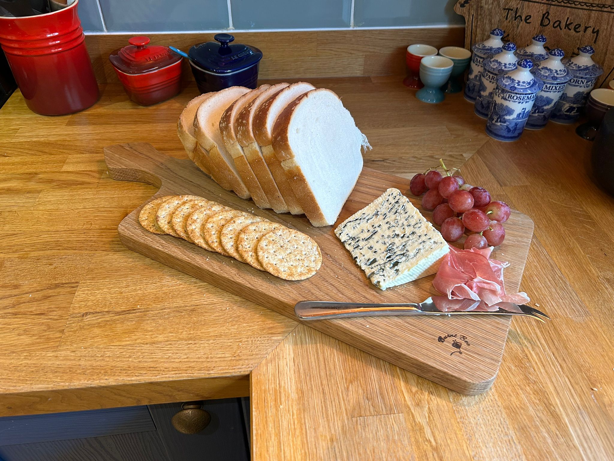 Picture of a solid oak paddle handled chopping board with bread, cheese, crackers, grapes and a knife on top of it